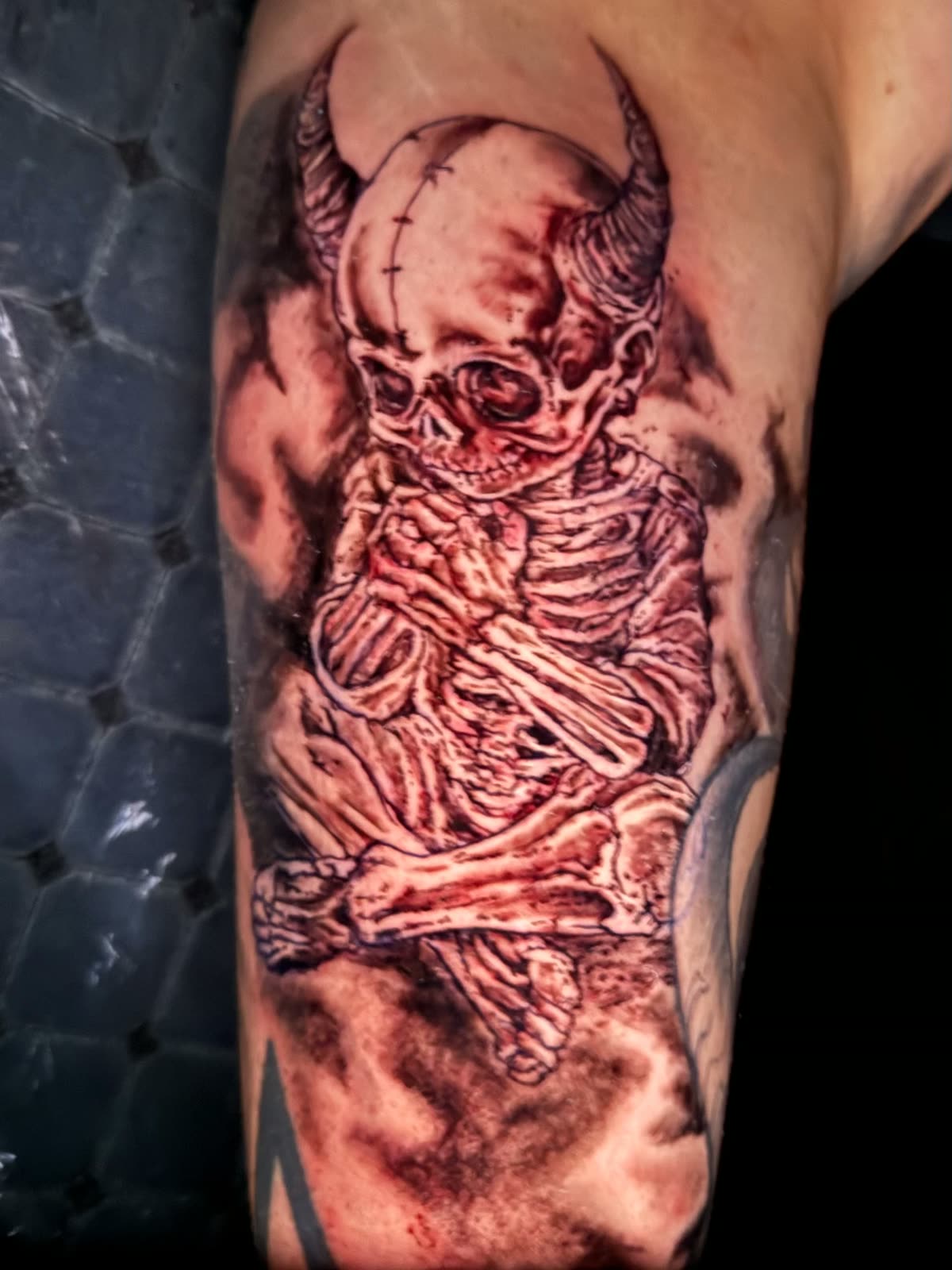 Black and grey horned skeleton tattoo seated with crossed legs on an upper arm