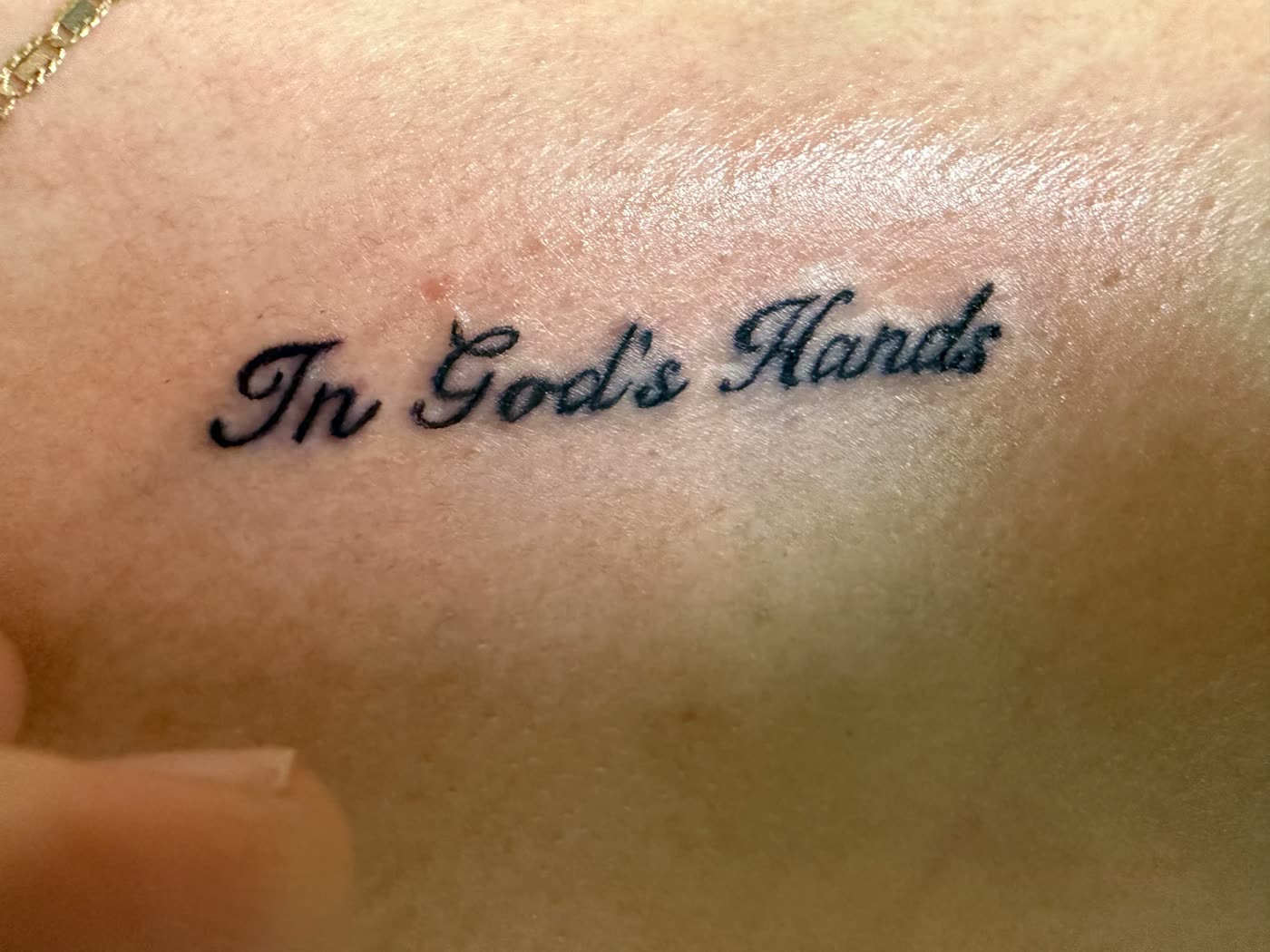 Fine-line black script tattoo reading In God's Hands on a collarbone